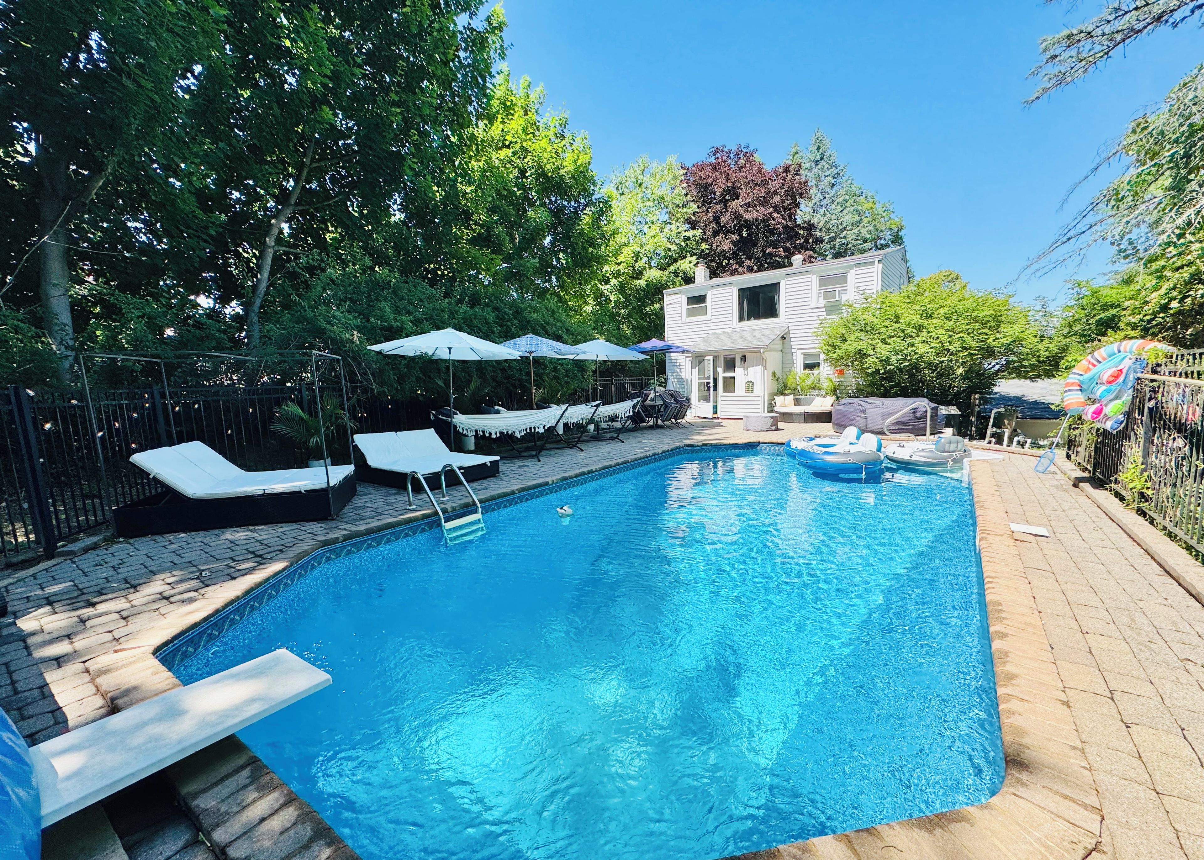 Una piscina rectangular con tobogán integrado, rodeada de sillas de descanso y sombrillas, con una casa y árboles al fondo.