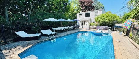 Una piscina rectangular con tobogán integrado, rodeada de sillas de descanso y sombrillas, con una casa y árboles al fondo.