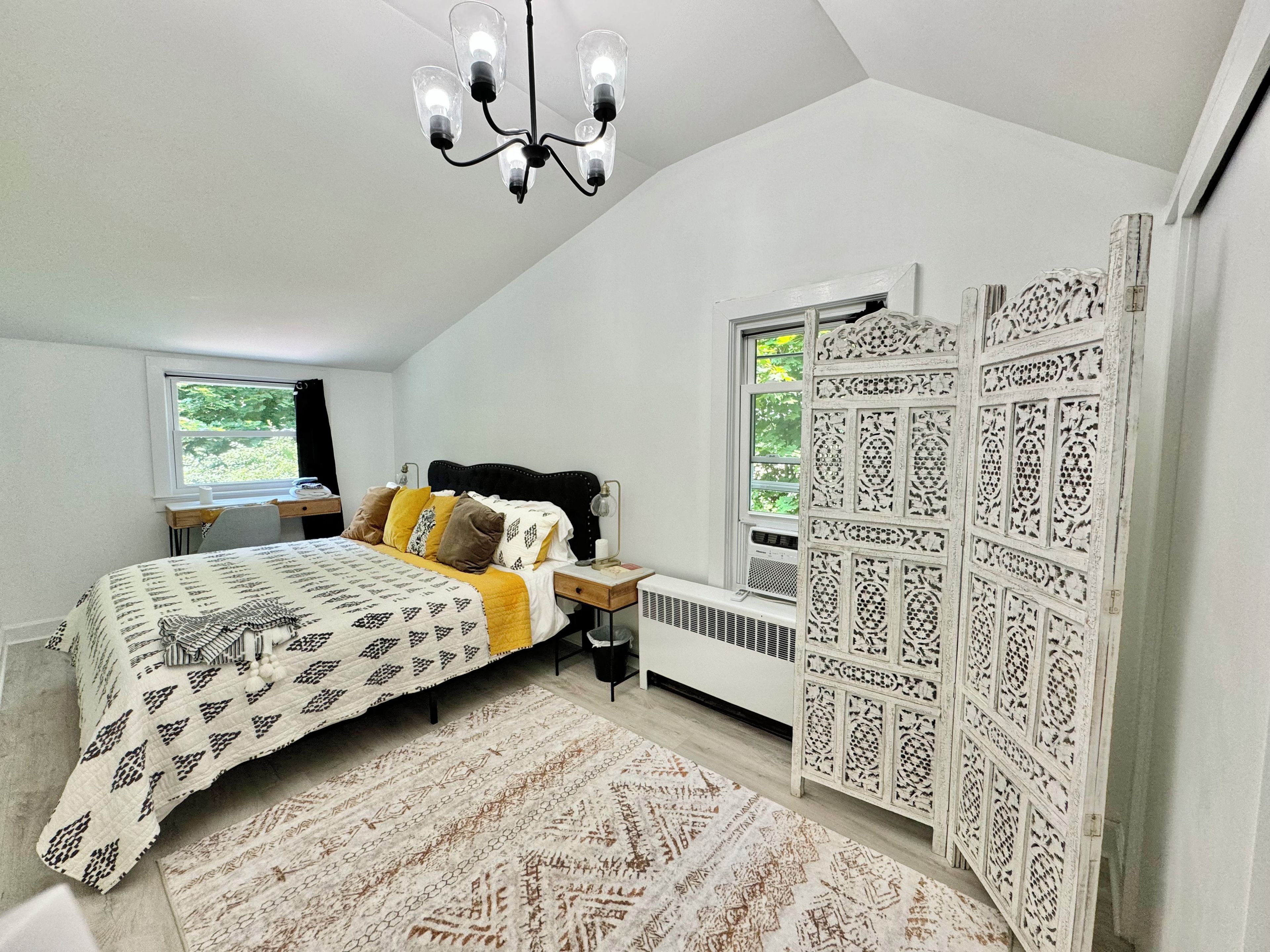 A bedroom with a bed, bedside table, lamp, radiator, window, and decorative panel.