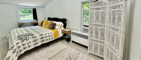 A bedroom with a bed, bedside table, lamp, radiator, window, and decorative panel.
