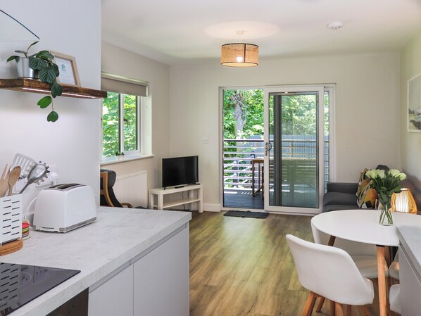 A modern kitchen with a toaster, a kettle, and a sink. There is a dining area with a table and chairs, and a balcony with a view of trees.