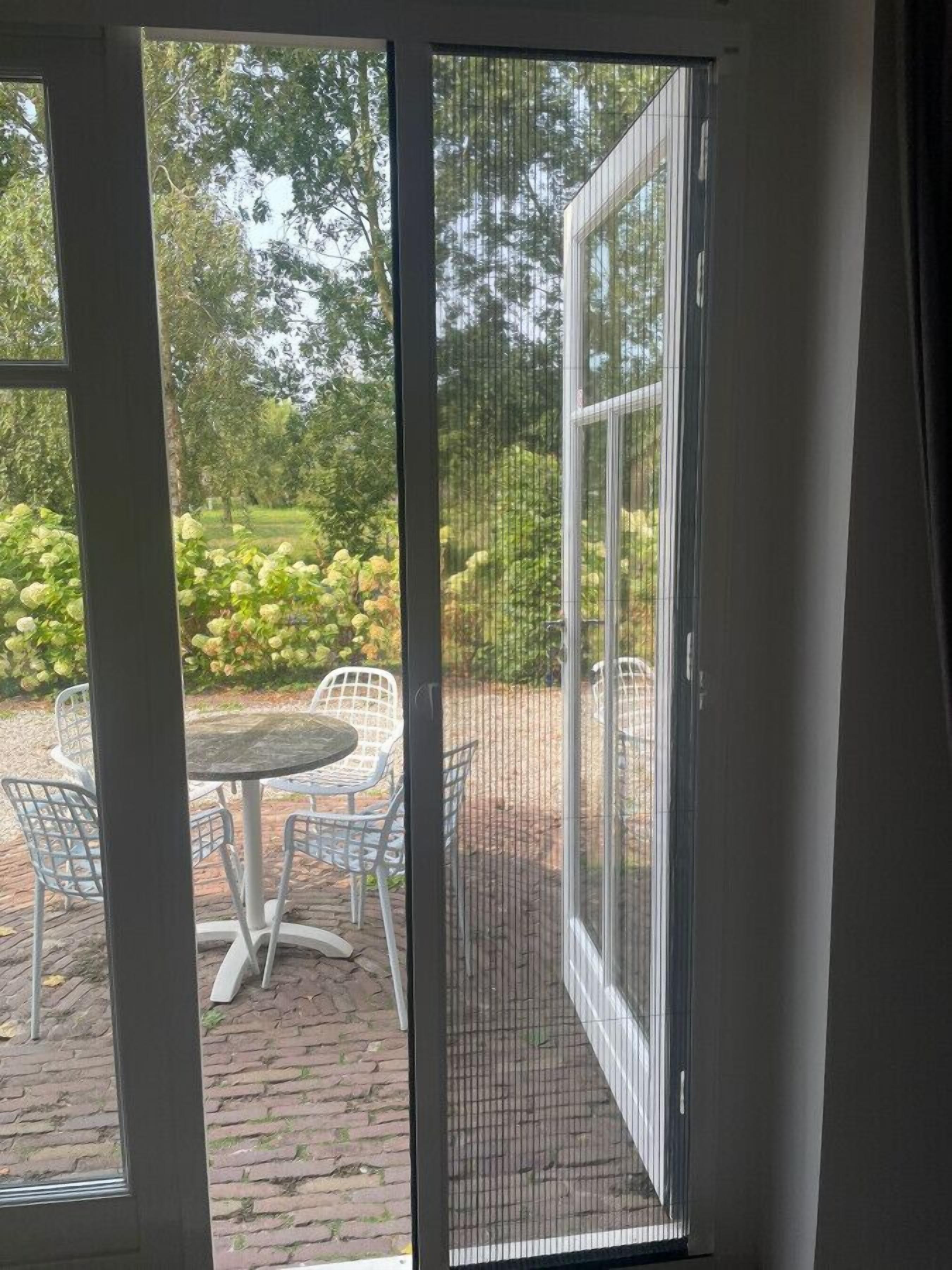Een terras met een tafel en stoelen, een tegelvloer en uitzicht op een tuin met bomen en struiken.