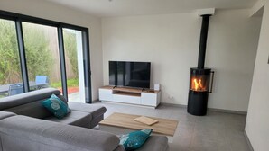 A modern living room with a grey sofa, a flat-screen TV, a wood-burning stove, and a large window overlooking a garden.