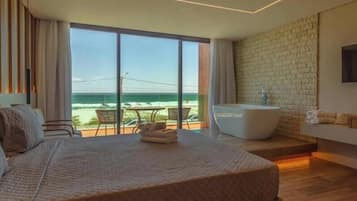 A modern hotel room with a large bed, a bathtub, a sofa, and a balcony with a view of the ocean.