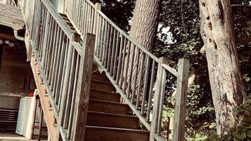 Eine Holztreppe führt zu einer Veranda mit einem Metallgeländer, umgeben von Bäumen und einem Kiesweg.