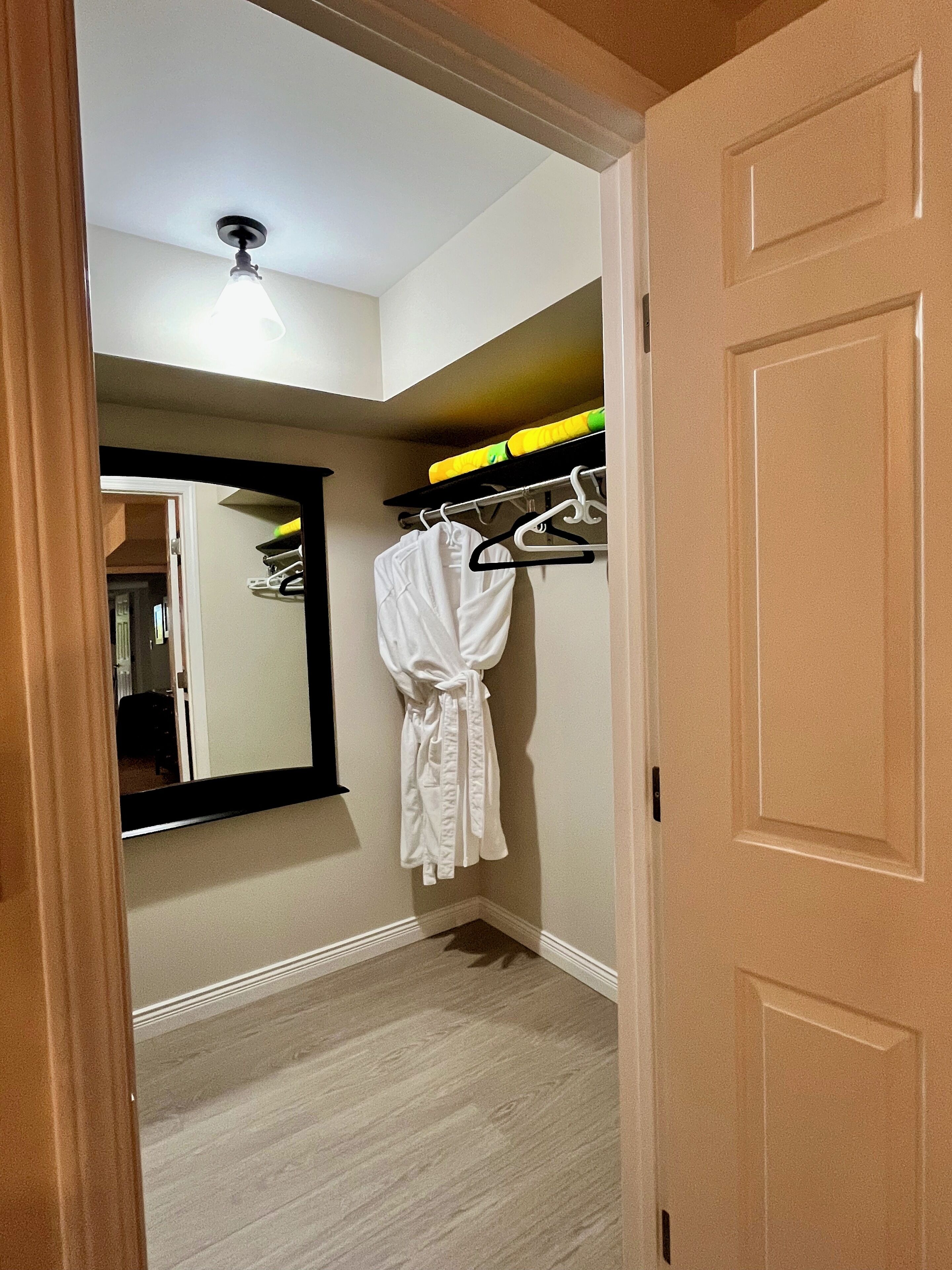 A hallway with a white robe hanging on a rack, a mirror on the wall, and a door leading to another room.