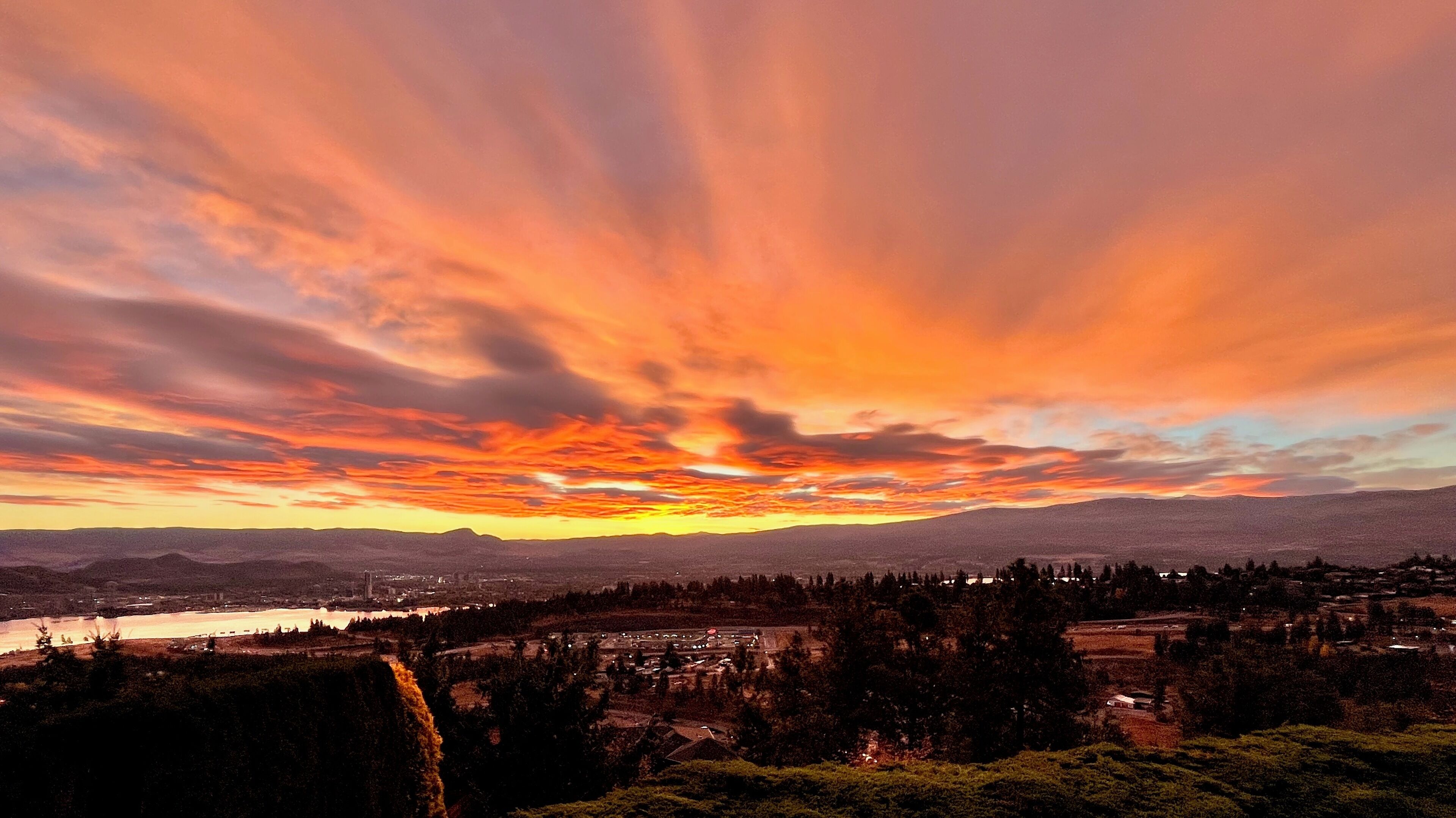 Ein weiter Panoramablick auf eine Stadt bei Sonnenuntergang, mit einem Fluss, Bergen und einem dichten Wald im Vordergrund.