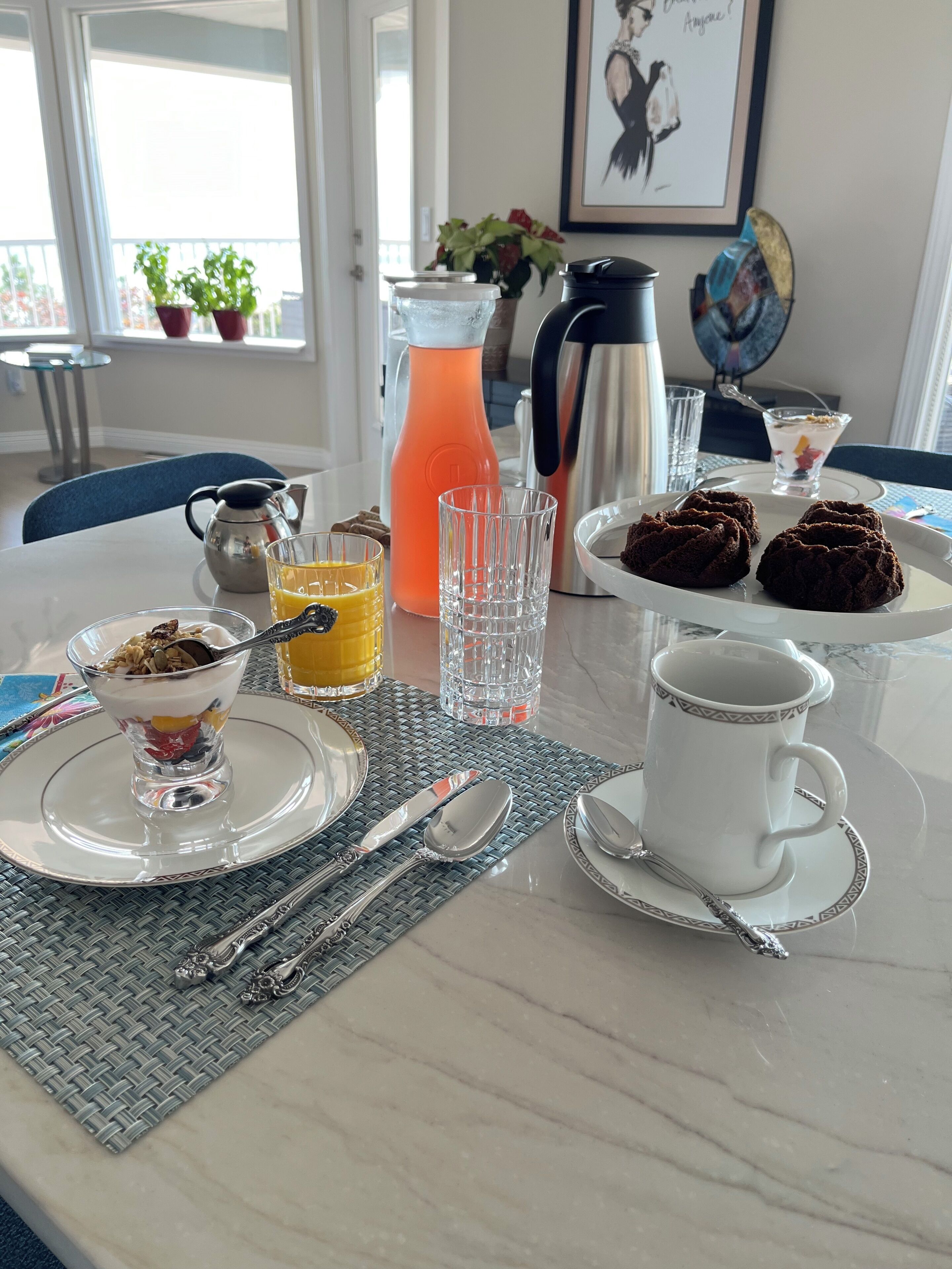 Una mesa de desayuno con una cafetera, un jarro, un vaso de jugo de naranja, un vaso de agua, un plato de galletas, una taza de café y un bol de parfait de yogur.