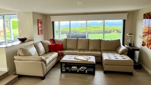 Un salon spacieux avec un grand canapé d’angle, une table basse et une vue sur un paysage verdoyant grâce à de grandes fenêtres.