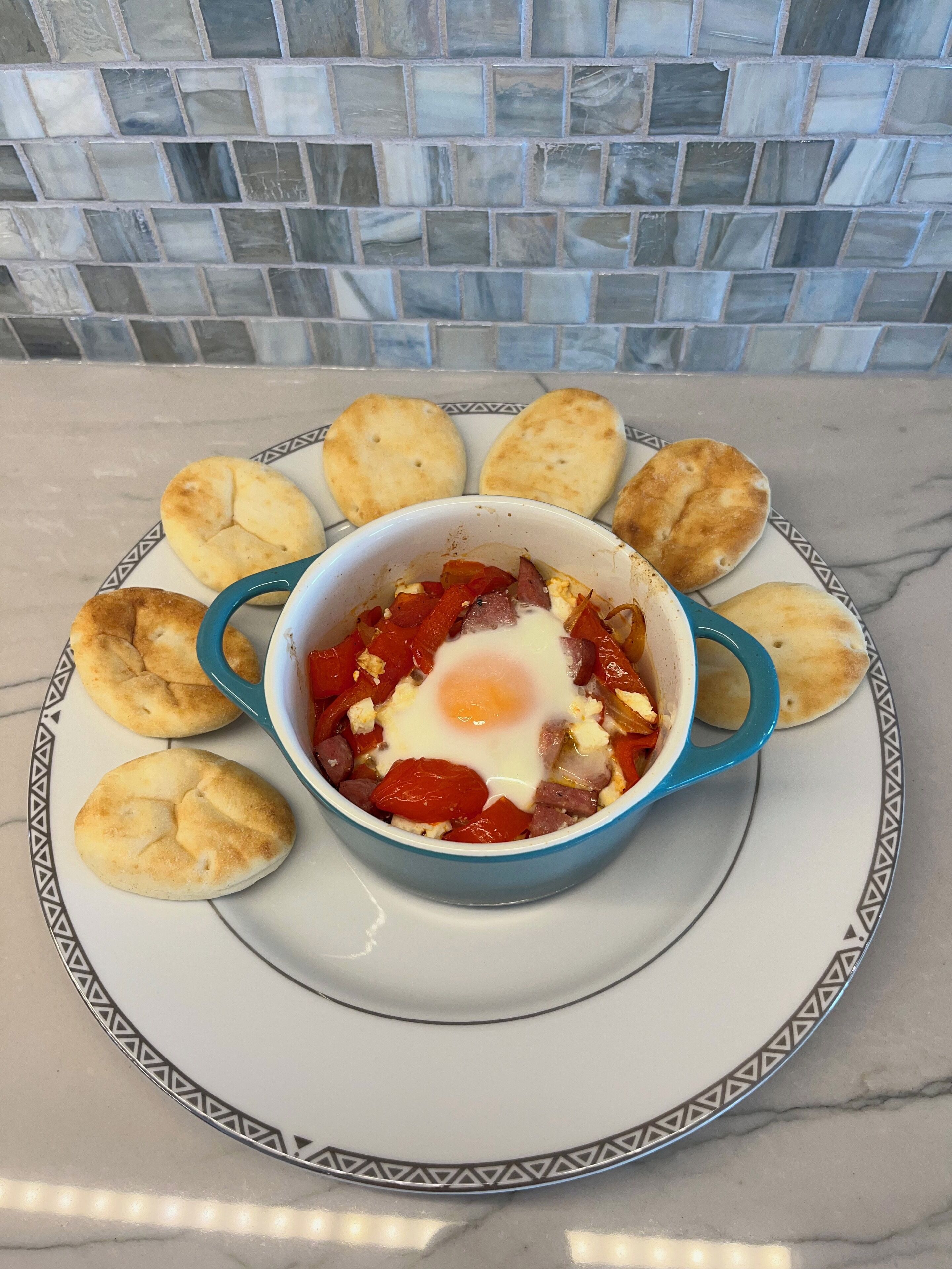 Prato de pequeno-almoço com ovos e legumes numa tigela azul, rodeado por pequenos pães num prato estampado.