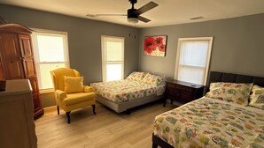 Un dormitorio con una cama grande, dos camas individuales, un sillón amarillo, un aparador de madera y un ventilador de techo.
