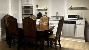Un comedor con mesa y sillas de madera, una cocina con armarios blancos, microondas y cafetera.