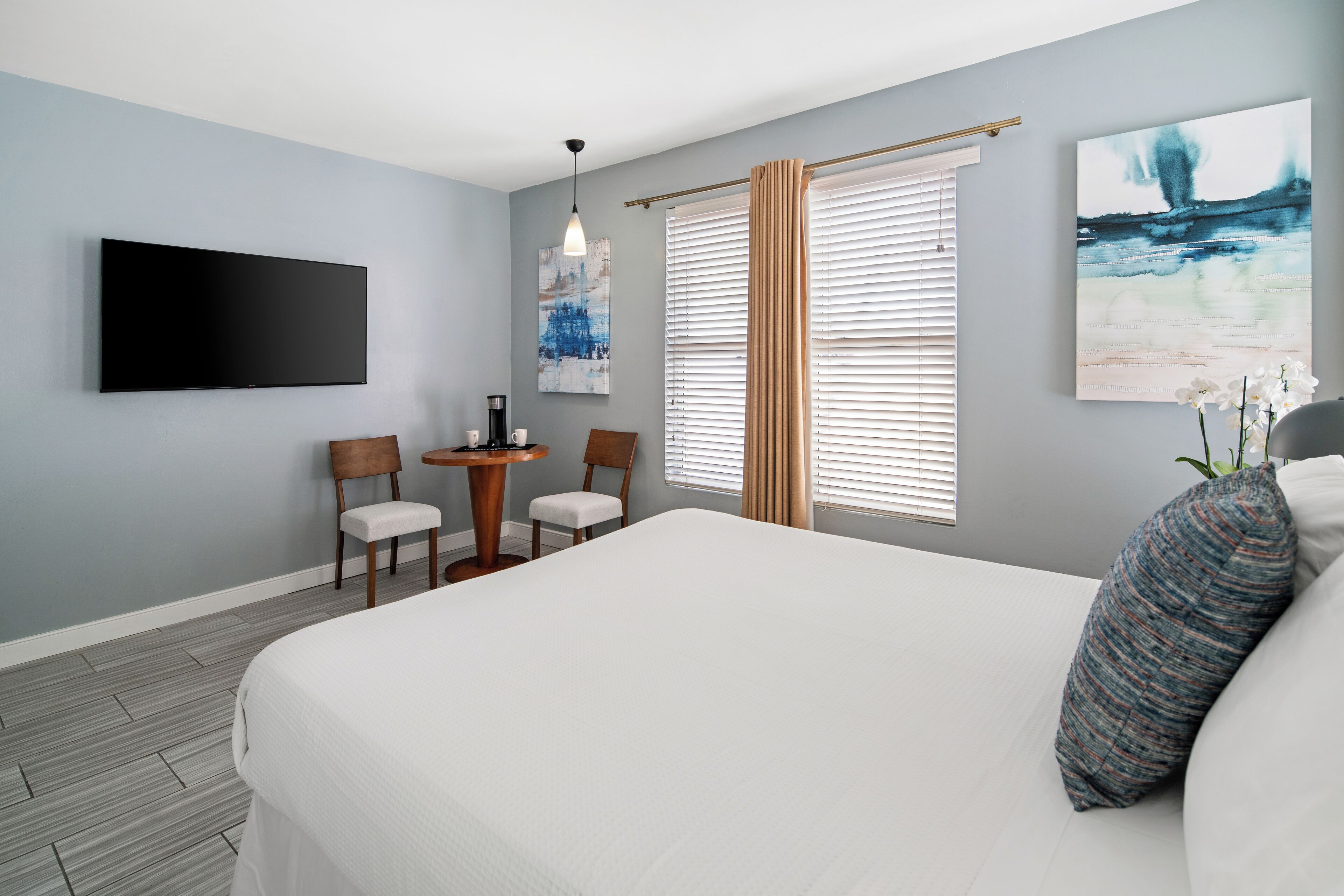 Ein Hotelzimmer mit einem großen Bett, einem flachbildfernseher an der Wand, einem kleinen Tisch mit Stühlen und Kunstwerken an den Wänden.