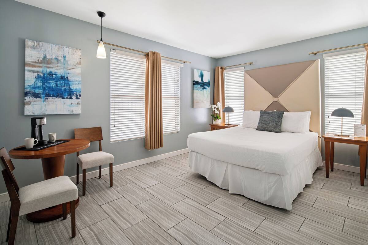 A modern hotel room with a large bed, a dining table, and two chairs. There are two windows with curtains, a painting on the wall, and a lamp on the bedside table.