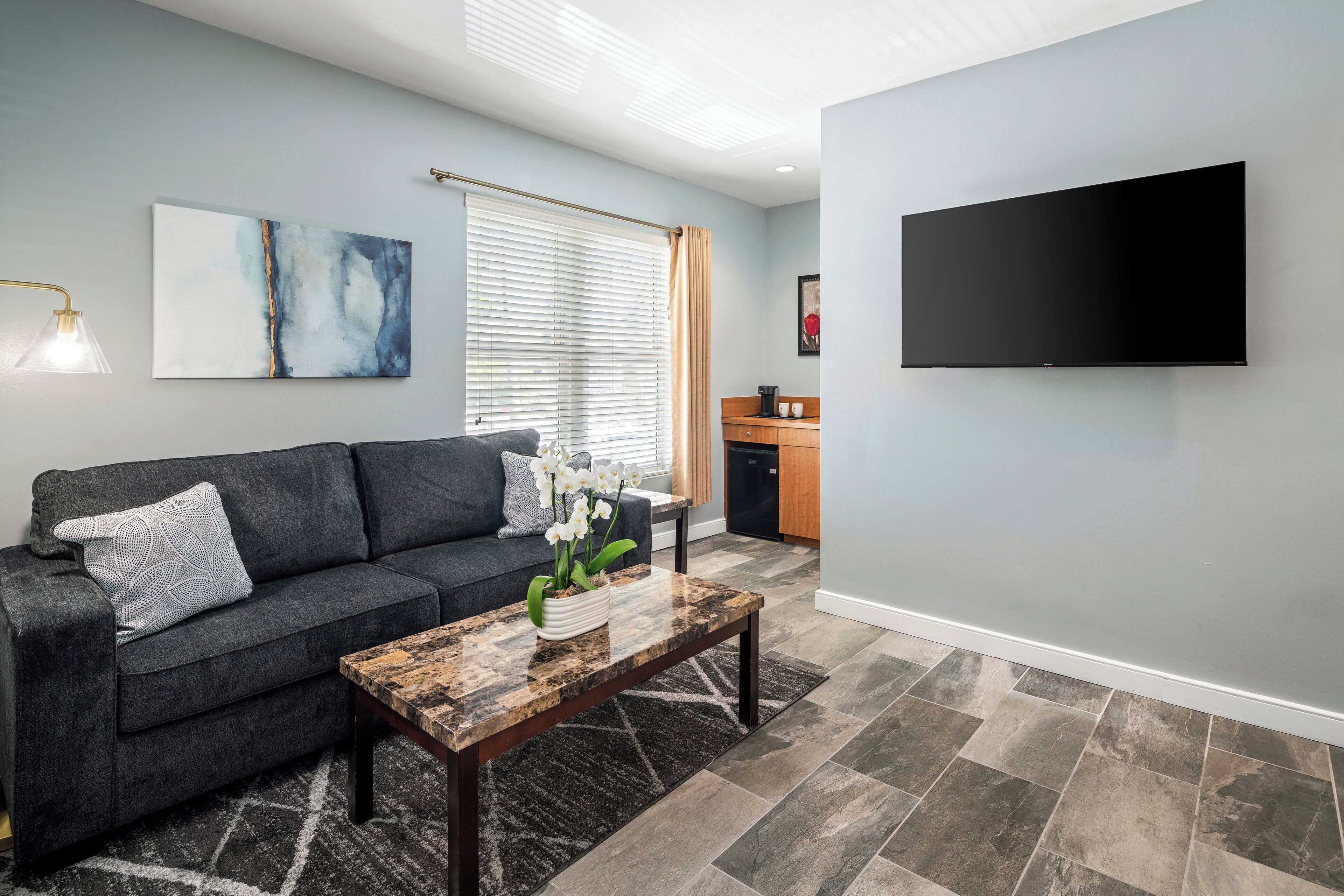 A modern living room with a dark grey sofa, a wooden coffee table with a vase of white flowers, a large abstract painting, a flat-screen TV mounted on the wall, and a kitchen area in the background.