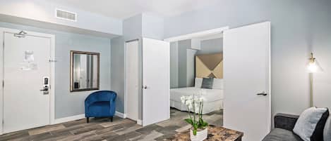 A modern hotel room with a check-in counter, a seating area with a sofa and armchair, a small table with a vase of flowers, and a bed with a headboard.