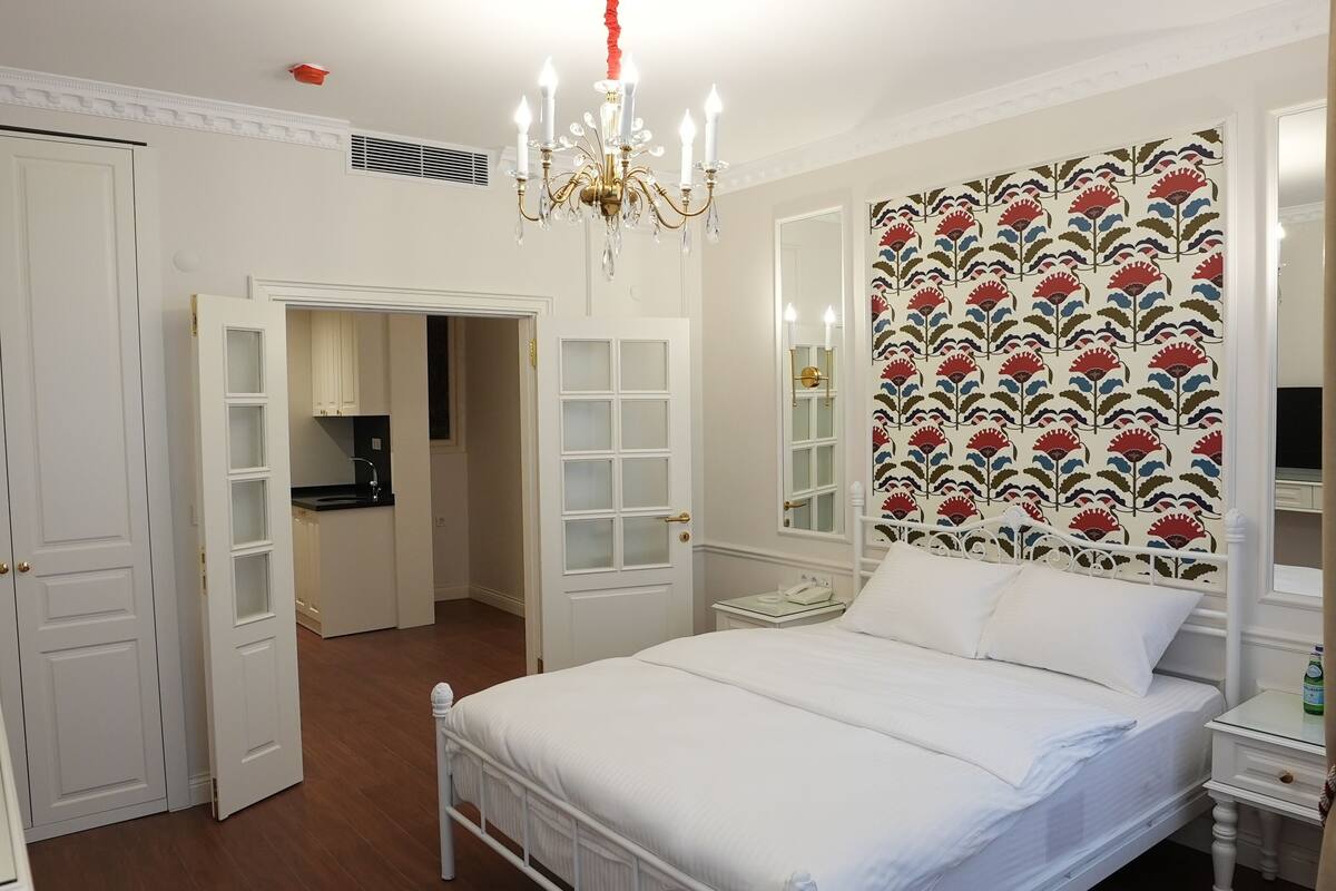 Ein Schlafzimmer mit einem weißen Bett, einer Chandelier, Tapete mit Muster und einem kleinen Nachttisch.