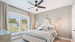 Une chambre à coucher avec un grand lit, un ventilateur de plafond, une fenêtre avec des stores et une table de chevet avec une lampe.