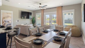 Un espace repas moderne avec une longue table dressée pour six personnes, un coin cuisine doté d’un four encastré et un coin salon confortable avec un fauteuil en forme de sac.