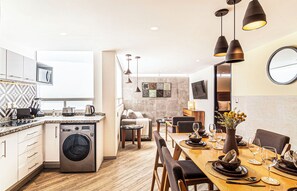 Eine moderne Küche mit eingebauter Waschmaschine, ein Essbereich mit einem Holztisch, der für eine Mahlzeit gedeckt ist, und ein Wohnbereich mit einer Couch und einem Fernseher.