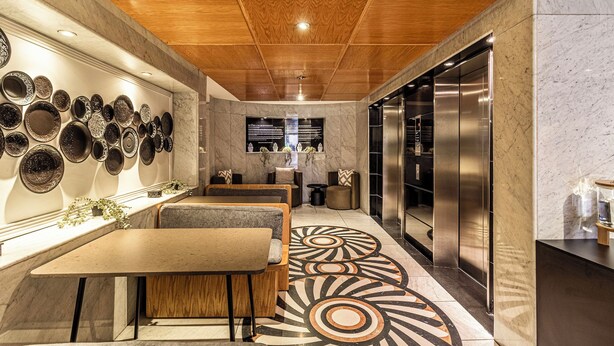 A modern interior with a wooden ceiling, a wall of decorative plates, a seating area, and a large elevator. - Kukun Grand Chapultepec (Mexico City)