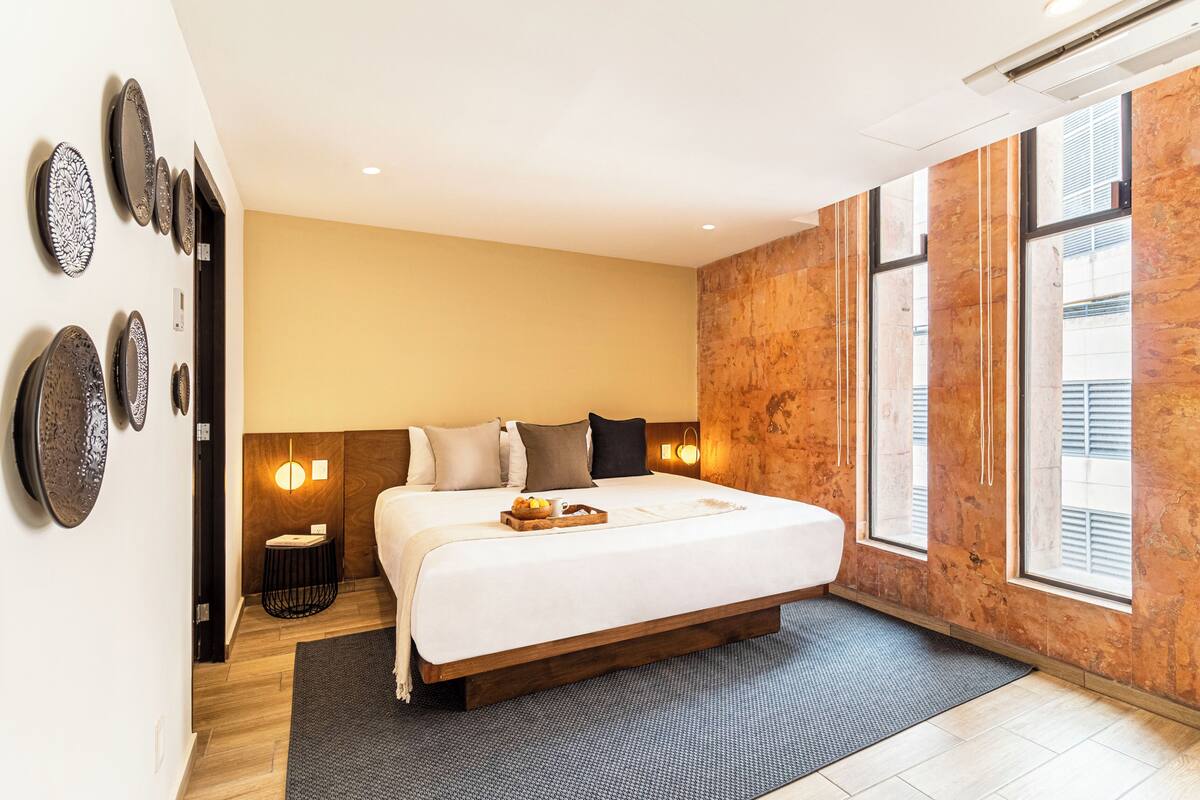 Habitación de hotel moderna con una cama grande, una mesita auxiliar, una lámpara de pared y un ventanal con persianas.