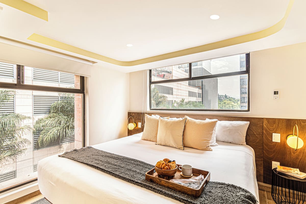 Una habitación de hotel moderna con una cama grande, un ventanal con vista a la ciudad, una bandeja con productos para el desayuno y una mesita de noche con lámpara.