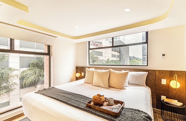 A modern hotel room with a large bed, a window with a city view, a tray with breakfast items, and a bedside table with a lamp.