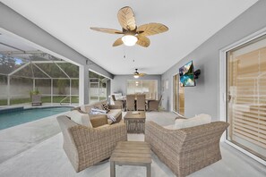 A spacious outdoor living area with a pool, wicker furniture, ceiling fans, and a TV mounted on the wall.