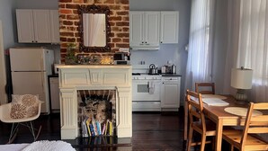 A cozy kitchen with a brick wall, white cabinets, a fireplace, and a dining area with wooden chairs and a table. - Cozy studio in enchanting New Orleans Neighborhood with AC, WiFi (New Orleans)