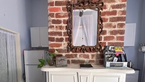 A room with a brick wall, a white fireplace, and a decorative mirror. There is a white desk with a plant and a box labeled "SWAMP HOUSE MAJESTIC REMOTES". - Cozy studio in enchanting New Orleans Neighborhood with AC, WiFi (New Orleans)