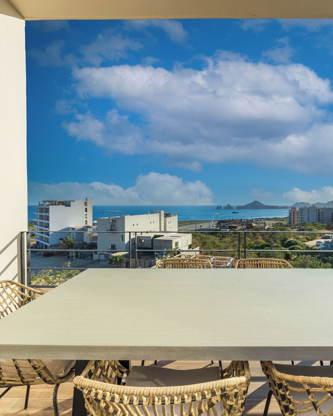 A balcony with wicker chairs and a table overlooking a coastal cityscape with buildings and a clear sky.