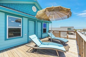 Un bâtiment turquoise doté d’une terrasse en bois, de deux chaises longues et d’un parasol de plage.