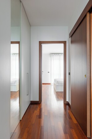 A hallway with wooden flooring, white walls, and a door leading to a bedroom with a bed and curtains.