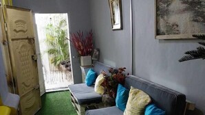 A room with a grey wall, a wooden door, a grey sofa with blue and yellow cushions, a small table with a vase of flowers, and a painting on the wall. - Miss. Emily Place (Monrovia)