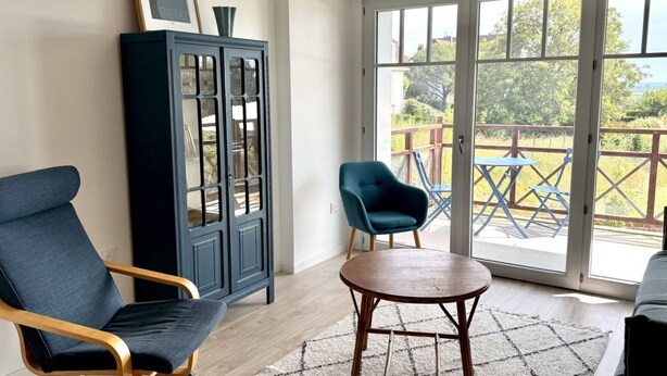 A room with a dark blue chair, a wooden cabinet, a small round table, and a view through a glass door to an outdoor area with a wooden deck and blue chairs.