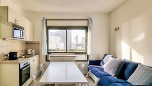A compact living space with a kitchenette, a dining table, a sofa, and a large window offering a city view. - Ben Yehouda Obsidian (Tel Aviv)