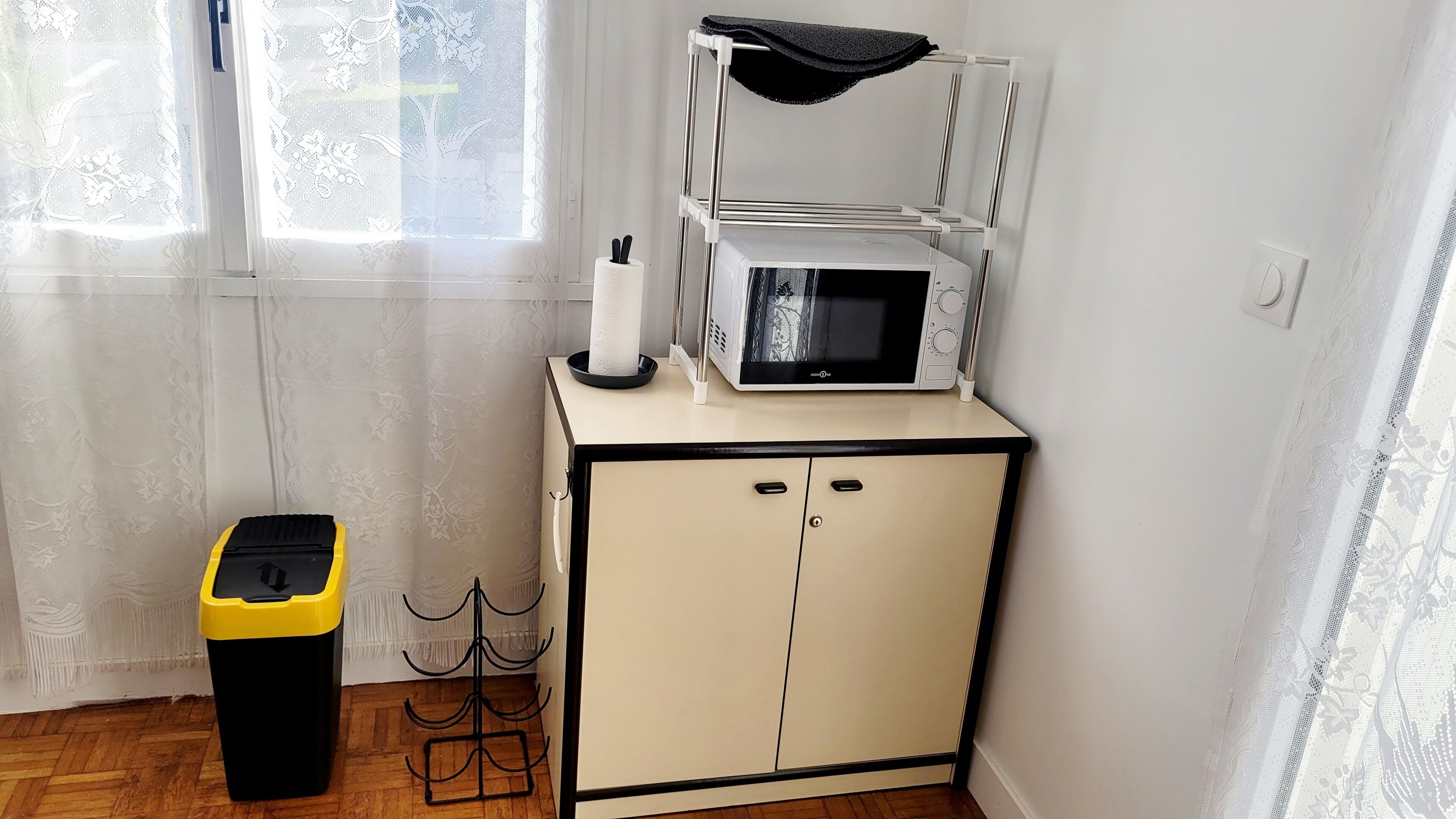 Een kitchenette met een magnetron, een klein kastje, een prullenbak en een raam met kantgordijnen.