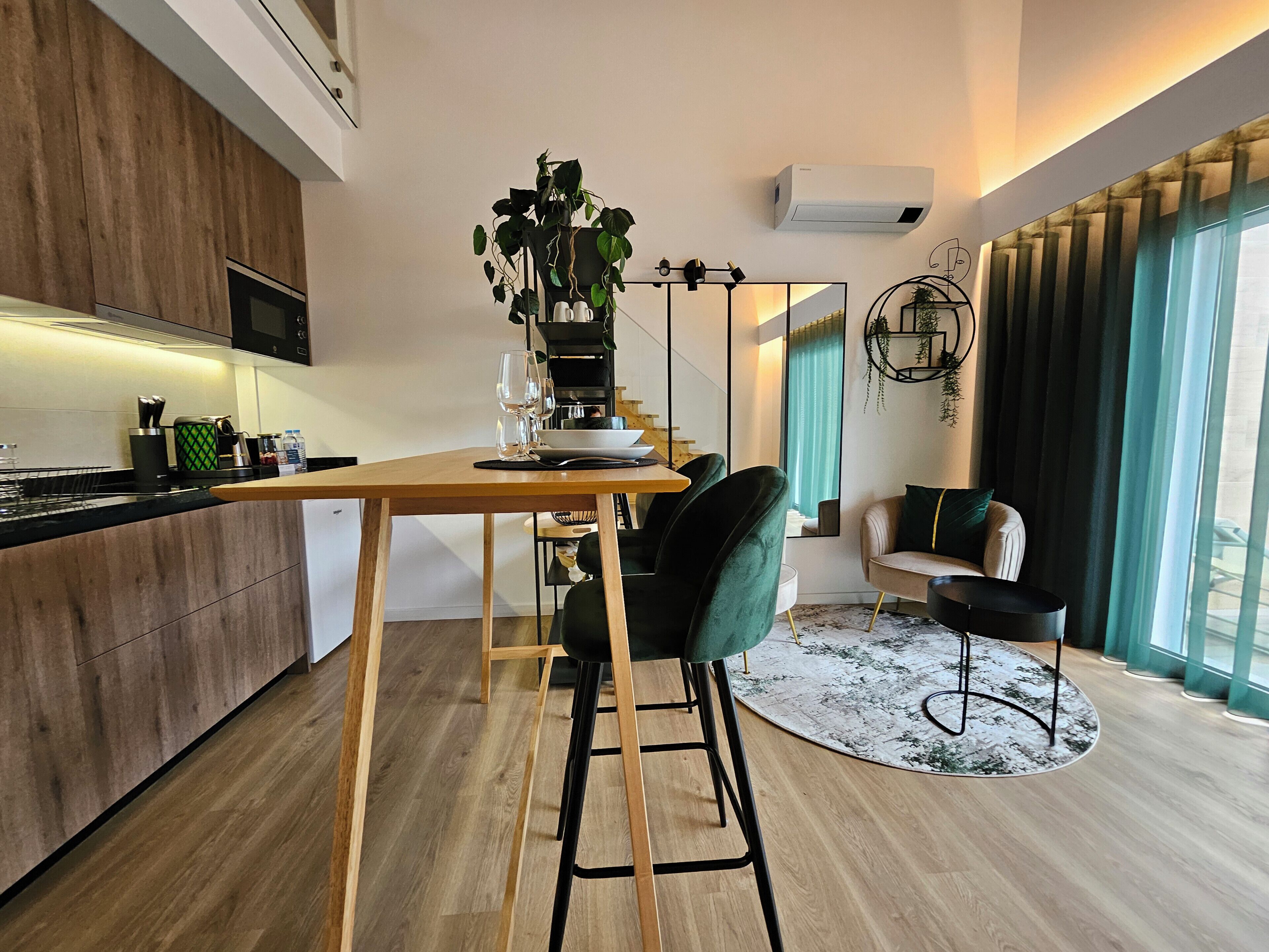 Une cuisine moderne avec des meubles en bois, un coin repas avec une table en bois et des chaises vertes, et un salon avec un canapé et une grande fenêtre.