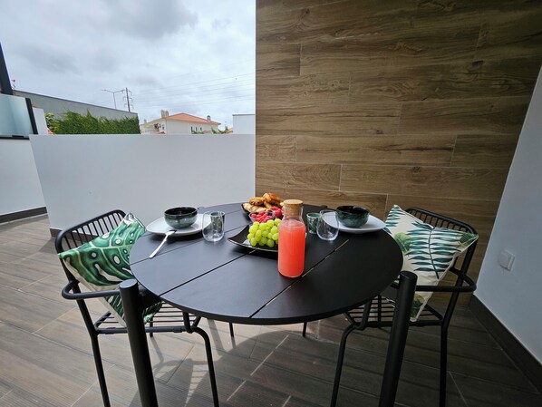 Un tavolo rotondo con sedie, una bottiglia di succo e un piatto di frutta su una terrazza affacciata su case e un cielo nuvoloso.