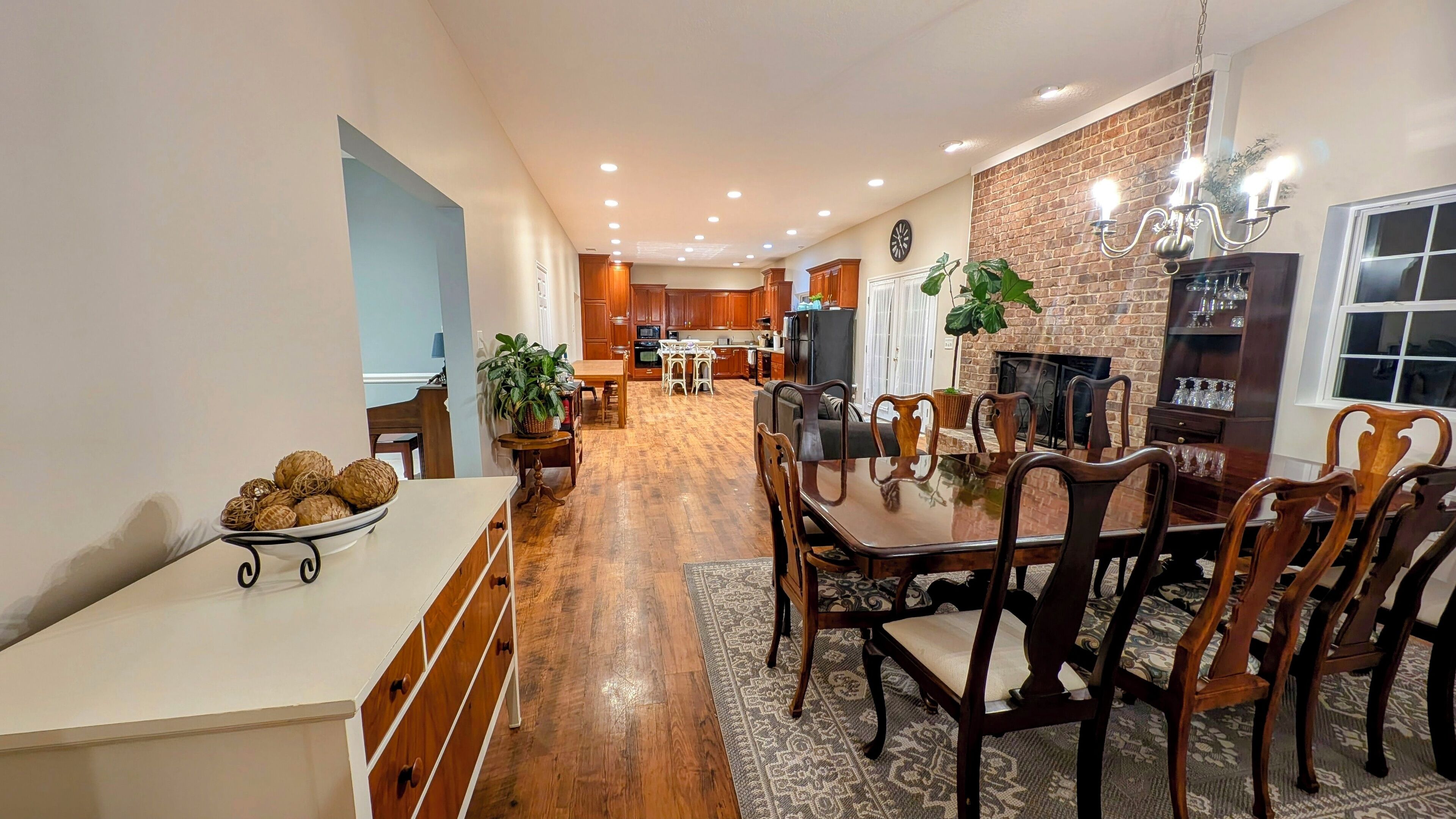 Una cocina amplia con armarios de madera, un comedor con mesa de vidrio y sillas de madera, y una pared de ladrillo con chimenea.