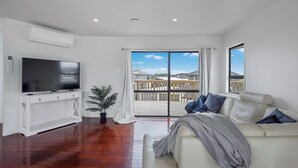 A modern living room with a flat-screen TV, a white sofa with cushions, a potted plant, and a sliding glass door leading to a balcony.