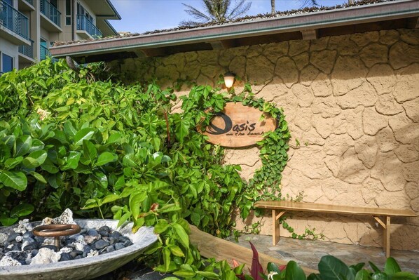 A stone wall with a wooden sign reading 'Oasis on the beach,' a wooden bench, and a fire pit with rocks.