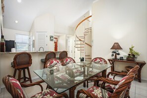 A dining area with a glass table surrounded by wicker chairs with floral cushions, a spiral staircase, and a small side table with a lamp and plant.