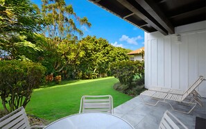 A patio with a table and chairs, a lawn, and a white building in the background. - Kiahuna Plantation Resort 403 Garden View 1 BD (Koloa)
