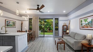 A modern living room with a sofa, coffee table, and a dining area with chairs and a table. There is a ceiling fan, recessed lighting, and a view through a glass door to a garden.