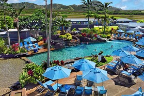 Área de piscina en un resort con múltiples sillas de descanso y sombrillas, rodeada de exuberante vegetación y vistas a montañas distantes.