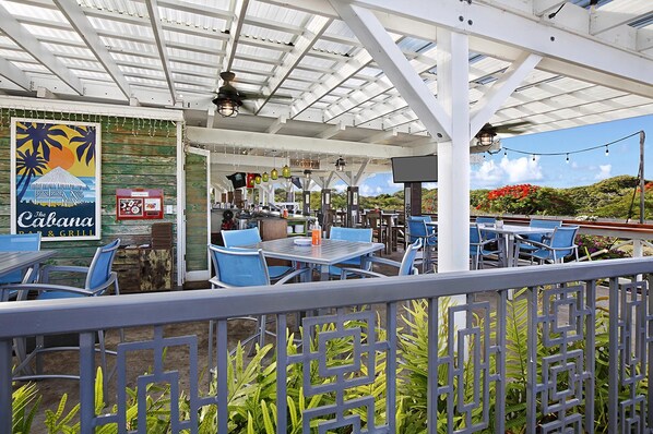 Un área de comedor al aire libre con sillas y mesas azules, un letrero que dice 'Cabana Bar & Grill', y vistas de un paisaje exuberante con flores rojas.