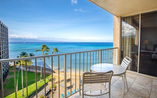 A balcony with a table and chairs, overlooking a beach with palm trees and the ocean.