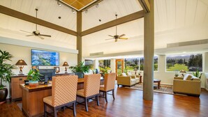 A spacious living room with a wooden floor, a dining area with patterned chairs, a large flat-screen TV, and a view of a green landscape through large windows.